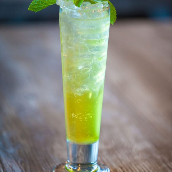 Tall glass of vibrant green iced drink with mint garnish on a wooden table.