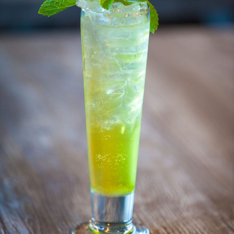 Tall glass of vibrant green iced drink with mint garnish on a wooden table.