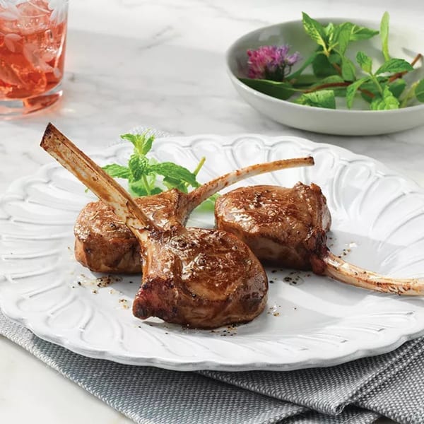 Three grilled lamb chops on a white plate with mint garnish, a drink, and a bowl of herbs.
