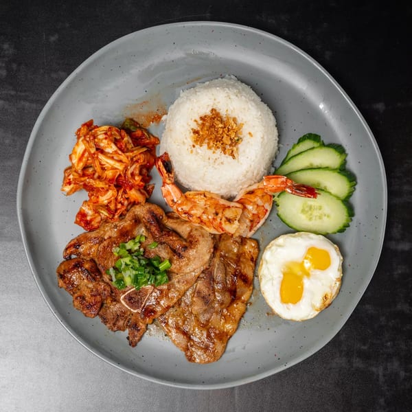 Grilled pork chops, shrimp, rice, kimchi, cucumbers, and fried eggs on a grey plate.