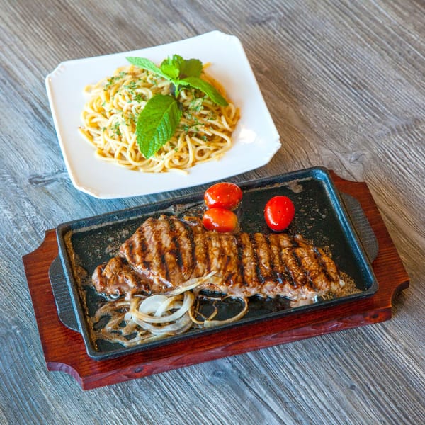 Sizzling steak with onions and tomatoes on a cast iron plate, next to a plate of spaghetti.